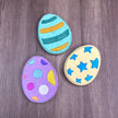 three easter egg bath bombs - one is yellow with blue stars one is purple with multi-color spots one is green with dark green & gold stripes