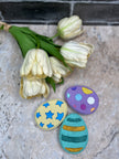 three easter egg bath bombs (green, purple & yellow) next to a bunch of ivory tulips on a tan background